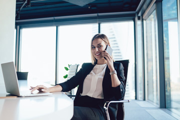 Wall Mural - Portrait of cheerful blonde woman entrepreneur in formal wear satisfied with good mobile phone conversation during working process,smiling female manager typing on laptop computer talking on cellular