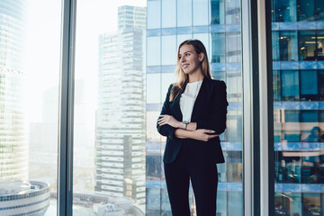 Wall Mural - Smiling blonde woman in trendy suit posing in office interior near window with panoramic view on business center, cheerful prosperous female proud ceo in formal outfit standing with crossed hands
