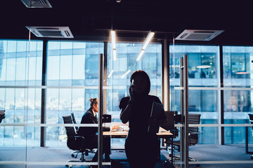 Wall Mural - Dark silhouette of businesswoman standing in shade of office interior talking on mobile phone about work, contour of female manager having smartphone conversation in dust working overtime on job