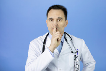 Wall Mural - Portrait of a Caucasian adult doctor in a white coat with a stethoscope over his neck on a blue background. Index finger at the mouth. Keep quiet. Close-up.