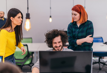 Two young businesswomen standing next to a male colleague while he is explaining new online tasks on a computer in a comfortable office atmosphere.