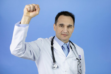 Wall Mural - Portrait of a Caucasian adult doctor in a white coat with a stethoscope over his neck on a blue background. Raised up hand clenched into a fist. Close-up. Soft focus. Face in blur