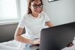 © nikkimeel - Young smiling woman uses laptop at home on the bed. Communication, video call, remote work