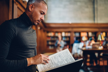 Wall Mural - Smart intellectual male professor reading book making researcher for work while standing in public library, concentrated adult student analyzing interesting information from literature manuals