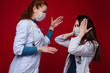 Side view of two doctors in white coats with stethoscopes arguing and gesturing. Brunette doctor covering her ears with hands.