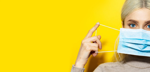 Wall Mural - Half face portrait of young blonde girl with blue eyes putting on medical flu mask on face, on yellow panoramic background with copy space.