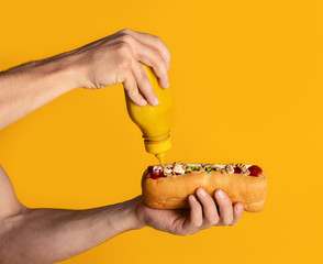 Wall Mural - Young man applying mustard on fresh juicy hotdog, orange background. Closeup