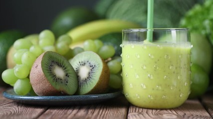 Wall Mural - Green tasty thick smoothie drink with seeds pouring glass stirring mixing up wooden table dark blue background grapes kiwi banana avocado cabbage. Vegetables and healthy vegan vegetarian beverage