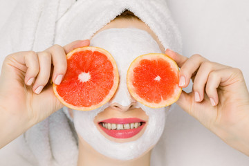 Wall Mural - Pretty young girl in bath towel and facial mask with grapefruit slices in front of her eyes. Beautiful woman holding grapefruit slices like eyeglasses, hiding her eyes, lying on white background