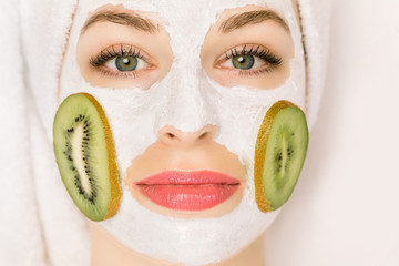 Wall Mural - Spa procedures and cosmetology. Close up top view of face of young attractive woman with fruit mask on her face woman and kiwi slices on cheeks, and with bath towel on head at spa saloon