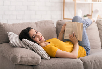 Wall Mural - Lockdown leisure activities. Young girl reading book while lying on sofa at home