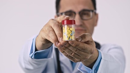 Wall Mural - Handsome middle aged doctor in medical coat, glasses and stethoscope shows pack of different colored pills on white background. Close up view focused on medicines, treatment, drugs poisoning concept