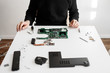 computer repairman sitting at a white table. on the table are different parts of a disassembled computer or laptop. Motherboard, cooling system, screws and screwdriver