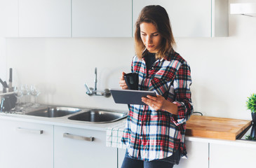 Wall Mural - Hipster girl using tablet technology and drink coffee in kitchen interior, person holding computer on background cuisine, female hands texting message, social distance lifestyle