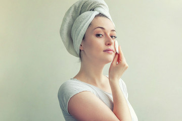 Wall Mural - Smiling woman in towel on head with soft healthy skin removing make up with cotton pad isolated on white background