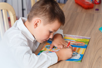 a seven year old boy in a white shirt diligently draws cars sitting at his Desk