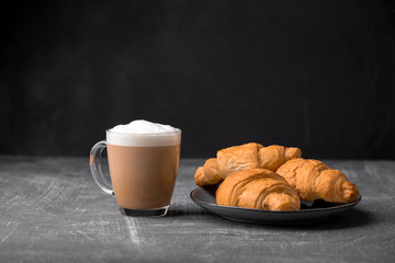 Wall Mural - Side view of delicious cappuccino coffee with milk foam sprinkled with croissants and spoon in a transparent glass mug on a gray background, horizontal format