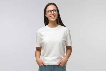 Happy student girl dressed in white T-shirt and jeans, wearing eyeglasses, looking aside with positive smile, isolated on gray background