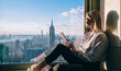 © BullRun - Side view of happy millennial digital nomad with modern touch pad looking in lookout window and dreaming about New York sightseeing, carefree hipster girl enjoying time in high rise building
