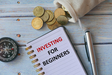 Top view of compass,gold coins,pen and notebook written with Investing for Beginners on wooden background.