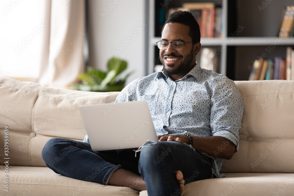 Smiling young african american man in eyewear relaxing on couch, looking at laptop screen. Happy multiracial guy web surfing internet, chatting with friends in social network, watching movie at home. - obrazy, fototapety, plakaty 