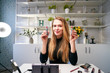 © artiemedvedev - Beauty blogger woman filming daily make-up routine tutorial near camera on tripod. Influencer blonde girl live streaming cosmetics product review in home studio. Vlogger female comparing lipsticks.