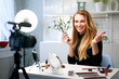 © artiemedvedev - Beauty blogger woman filming daily make-up routine tutorial near camera on tripod. Influencer blonde girl live streaming cosmetics product review in home studio. Vlogger female comparing lipsticks.