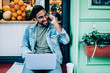 © BullRun - Young man speaking on cellphone and working on laptop next to fruit shop