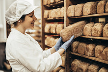 Young Female Worker Working In Bakery. She Puts Bread On Shelf.