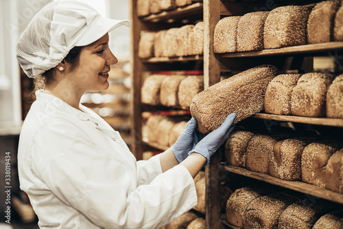 Fotografía Young female worker working in bakery. She puts bread on shelf.
