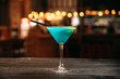 Side view on sweet delicious cocktail blue lagoon with a black straw in the martini glass on the wooden table with blurry bokeh lights background
