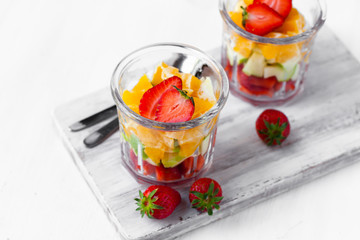 Concept of healthy food, clean eating. Low calories sweet summer dessert. Homemade simple fruit salad in two glasses for breakfast. Orange, apple, strawberry. White wooden background, copy space