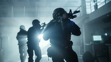 Masked Squad Of Armed SWAT Police Officers Move In A Hall Of A Dark Seized Office Building With Desks And Computers. Soldiers With Rifles And Flashlights Surveil And Cover Surroundings.