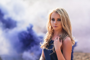 Young beautiful blond woman in blue mini dress and floral wreath standing and trying water on summer day with coutryside landscape at background. Woman natural beauty and summer nature concept. smoke