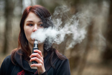 Vape Teenager. Young Pretty Caucasian Brunette Girl Smoking An Electronic Cigarette On The Street In The Spring. Deadly Bad Habit.