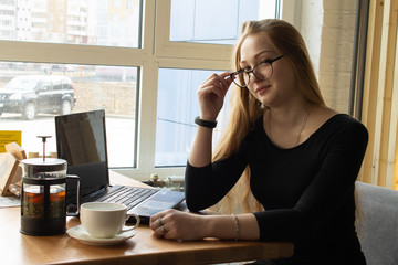 Beautiful sexy girl in a black dress and glasses sits at the table and works at the computer. Distance learning. Remote work.