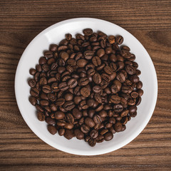 Coffee beans on a round white plate. Wooden brown background. Selective focus. Top view.