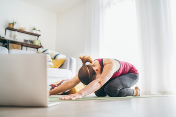 Fit sporty healthy woman on mat in Child’s Balasana gentle resting yoga pose, doing breathing exercises, watching online yoga class on laptop computer. Healthy people and self motivation concept.