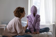 © fizkes - Stubborn teenage girl puts hood on head avoiding mom apology, ignoring talk with parent sitting on bed at home. Naughty teen daughter introvert hiding in hoodie showing disrespect to mother at home.