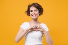 Smiling Young Brunette Woman Girl In White T-shirt Posing Isolated On Yellow Orange Background In Studio. People Lifestyle Concept. Mock Up Copy Space. Showing Shape Heart With Hands Heart-shape Sign.