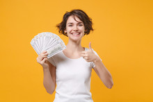 Smiling Young Brunette Woman In White T-shirt Posing Isolated On Yellow Orange Background. People Lifestyle Concept. Mock Up Copy Space. Hold Fan Of Cash Money In Dollar Banknotes Showing Thumb Up.