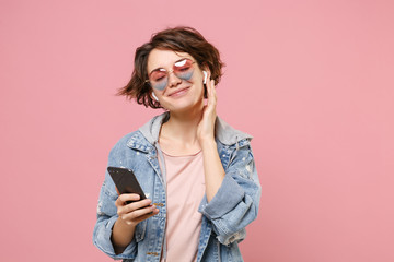 Wall Mural - Charming young brunette woman in casual denim jacket, eyeglasses posing isolated on pastel pink background. People lifestyle concept. Mock up copy space. Listen music with air pods, hold mobile phone.