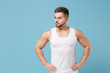 Attractive young bearded guy 20s in white singlet posing isolated on pastel blue background. Sport fitness healthy lifestyle concept. Mock up copy space. Stand with arms akimbo on waist looking aside.