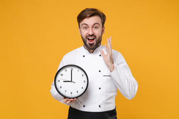 Wall Mural - Excited young bearded male chef cook or baker man in white uniform shirt posing isolated on yellow background studio portrait. Cooking food concept. Mock up copy space. Hold clock, spreading hands.