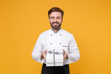 Wall Mural - Smiling young bearded male chef cook or baker man in white uniform shirt posing isolated on yellow wall background. Cooking food concept. Mock up copy space. Hold present box with gift ribbon bow.