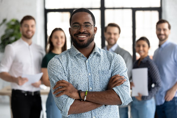 Smiling motivated African American male employee stand forefront posing together with colleagues in office, happy biracial businessman show success and leadership in business, employment concept