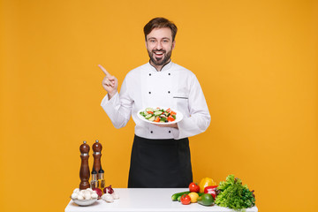 Wall Mural - Excited bearded male chef cook or baker man in white uniform isolated on yellow background. Cooking food concept. Mock up copy space. Stand near table hold plate with salad pointing finger aside up.