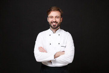 Wall Mural - Smiling young bearded male chef cook or baker man in white uniform shirt posing isolated on black wall background studio portrait. Cooking food concept. Mock up copy space. Holding hands crossed.