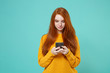 © ViDi Studio - Pretty young redhead woman girl in yellow sweater posing isolated on blue turquoise background studio portrait. People lifestyle concept. Mock up copy space. Using mobile phone, typing sms message.