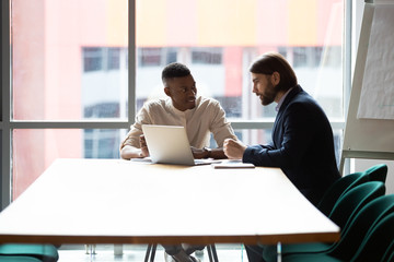 Smiling multiracial colleagues sit at office table in boardroom brainstorm using modern laptop together, multinational coworkers or partners talk discuss financial ideas work on computer at briefing